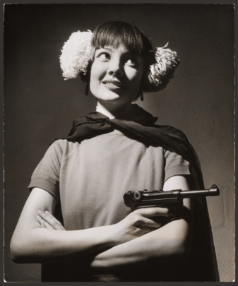 A white woman wearing earmuffs, a cape, and carrying a toy gun, poses with her arms crossed